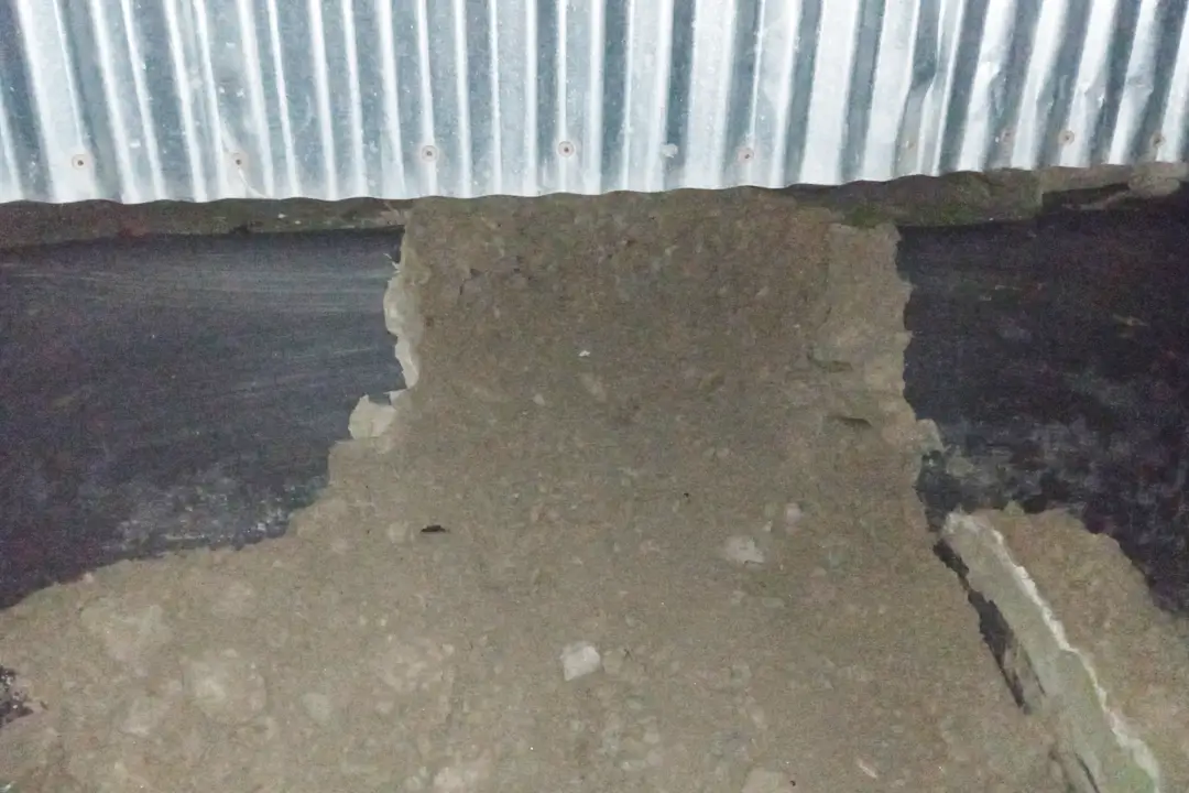 A rural Bangladeshi village scene at night, where locals holding flashlights are investigating a massive hole dug beneath the base of a traditional mud house, revealing the unusual method used by thieves during a robbery.
